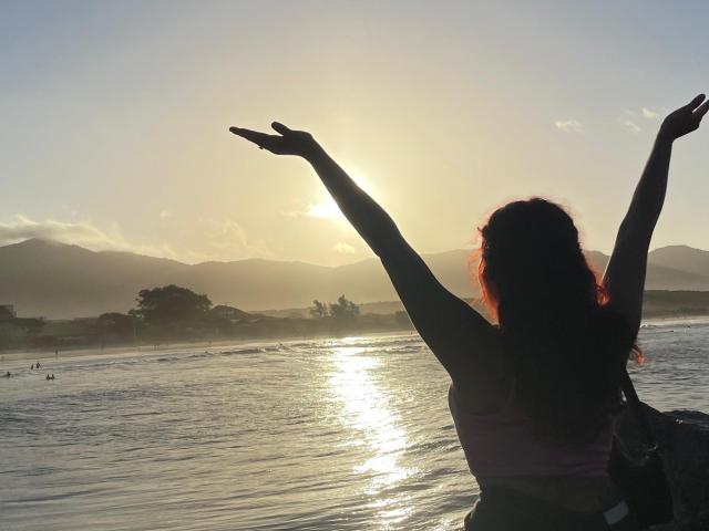 Silhouette of SweetSolei69 with arms raised, facing sunset over a reflective water surface. She has long red hair and is wearing a pink sports bra. Sunlight glistens on water.