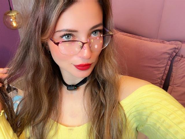 AmbeerWood has long brown hair, blue eyes, and red lipstick, wearing glasses with star patterns, a black choker, and a yellow off-shoulder top, showing her cute face.