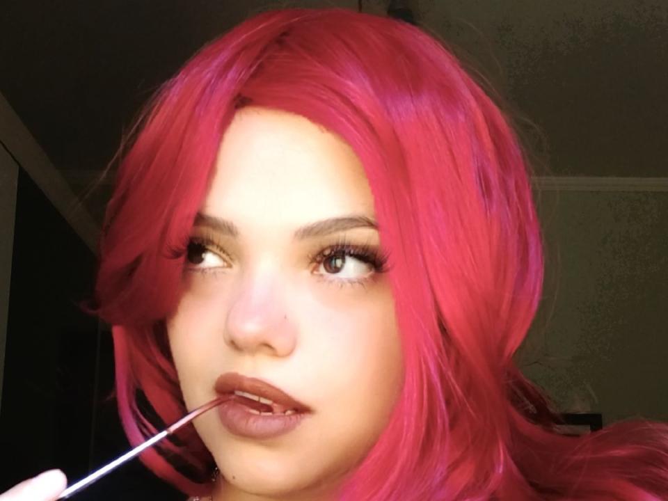 MahEla, com o cabelo rosa vibrante, lábios carnudos e um lápis na boca, olhando para o lado com uma expressão ligeiramente entediada, segurando o lápis perto do lábio superior..