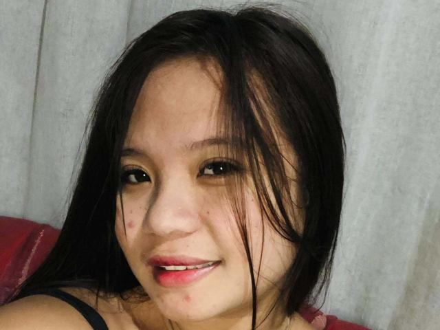 YannaDirtyOne has long black hair, a cute smile with slightly parted red lips, and small pimple-like blemishes on her face, wearing a black top, in a selfie.
