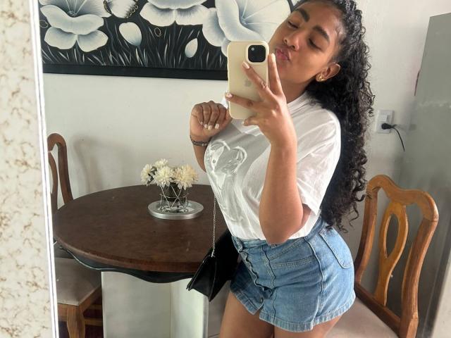 NatalyBlonder, avec ses cheveux noirs bouclés, portant un haut blanc transparent et un short en jean bleu taille haute, embrasse son téléphone tout en se prenant une photo devant le miroir, montrant ses fesses..