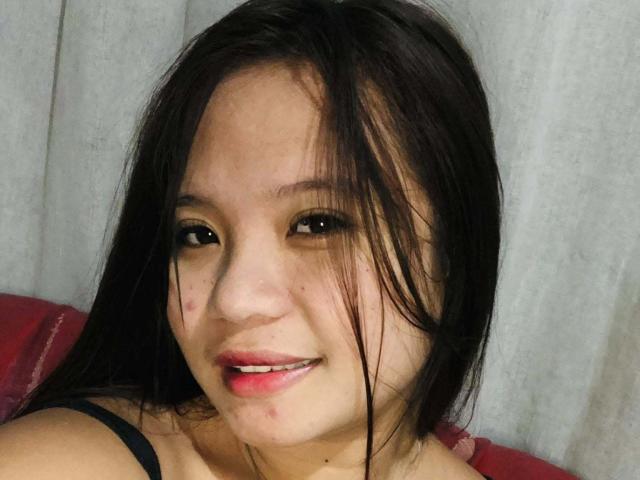 YannaDirtyOne with long black hair, slightly messy, smiling with pink lips, small zits on her face, black strap top, light skin, brown eyes, red pillow behind her.