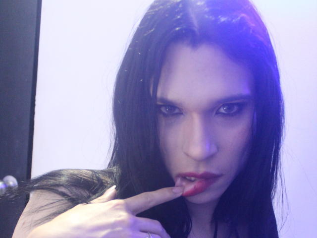 Jade Oconner, con cabello oscuro, ojos azules y lápiz labial rojo, chupándose el dedo, mirando seductoramente a la cámara, con una ligera luz púrpura-azul en su rostro..