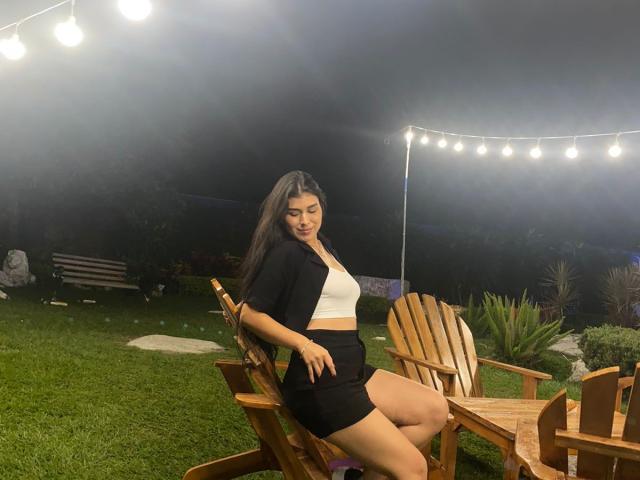 FernandaMl mit langem, dunklem Haar, weißem Crop-Top, schwarzer Jacke und schwarzem Rock, sitzend auf einem Holzstuhl, Nacht, Lichterketten, Gras und Holzstühle im Hintergrund..