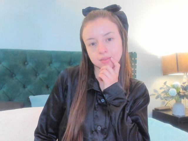 VioletaNova met lang bruin haar, een zwarte strik en een zwart shirt, die haar vinger bijt, ziet er schattig uit, zittend in een bed met een groene bekleding, naast een lamp en een bloevas..