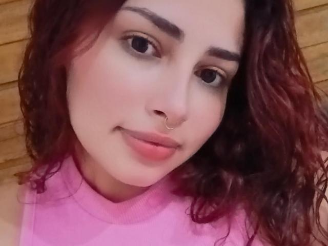 SweetSolei69 tiene el pelo castaño rizado, ojos marrones y un pequeño piercing en la nariz. Lleva una blusa rosa y sonríe suavemente, con los labios rosados. Sus senos no son visibles..
