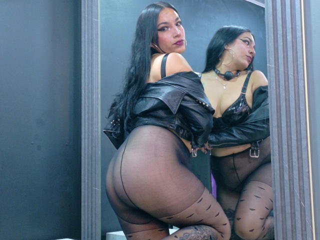 KiaraThomsomp with long black hair, wearing a black leather jacket, sheer black pantyhose, and a black bra, showing her ass in a mirror reflection. Her tits are visible.