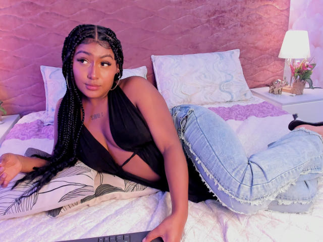 AmberrQueen lying on a bed, wearing a black halter top that shows her tits, and blue ripped jeans. She has long braids, hoop earrings, and a tattoo on her chest.