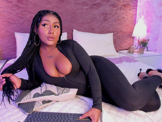 AmberrQueen with big tits, black long-sleeve zip-up, laying on bed, braids, holding keyboard, pillow under arm, white sheets, pink wall, lamp, and flower vase in background.
