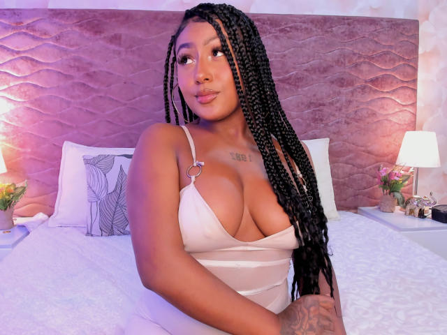 AmberrQueen with large tits, wearing a white, low-cut, strapless dress, has long black braids, brown skin, and a tattoo above her cleavage, sitting on a bed.