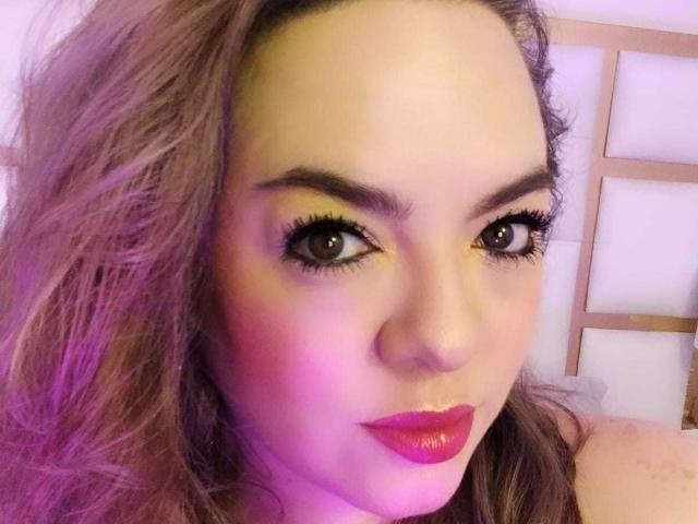 AvaMia's face, with pinkish-purple hair, thick eyelashes, and shiny pink lipstick, staring at the camera with brown eyes and slightly arched eyebrows, in a soft purple light.
