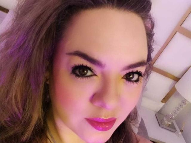 AvaMia has pink lipstick, purple eyeshadow, and long eyelashes. Her brown hair is loose, and she has a slight smile with purple lighting on her face.