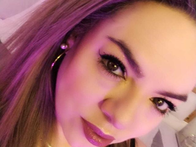 AvaMia has purple lipstick, long eyelashes, and purple-tinted hair with matching earrings, looking at the camera with a slight smile, her eyes are brown and she's wearing purple eyeshadow.
