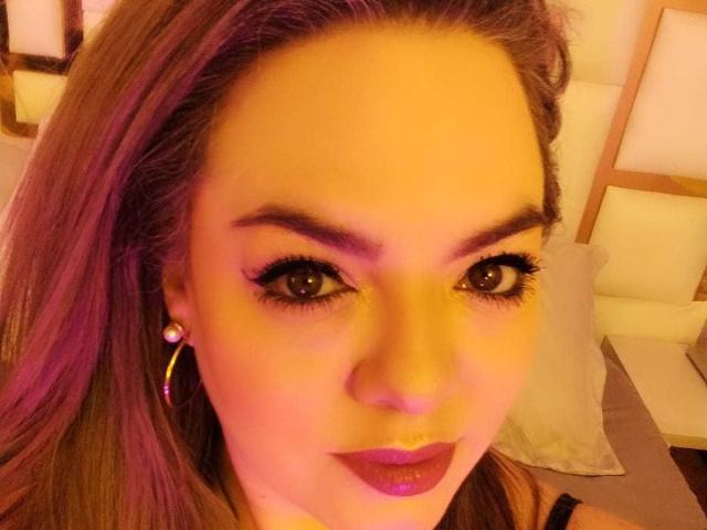 AvaMia's face with purple lipstick, hoop earrings, and pink highlights in her hair, glowing yellow under warm lighting, brown eyes with long eyelashes, and a slight smile.