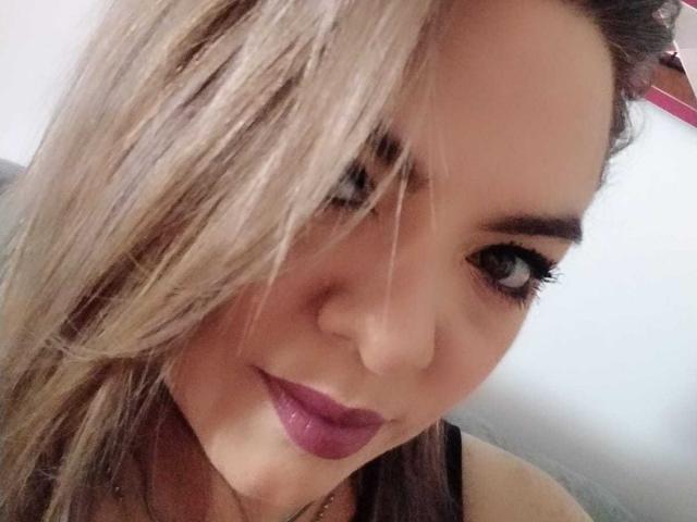 AvaMia's face, blonde hair covering one eye, dark purple lipstick, slight smile, black tank top, visible eyelashes, white background, close-up selfie, soft lighting, slightly messy hair.