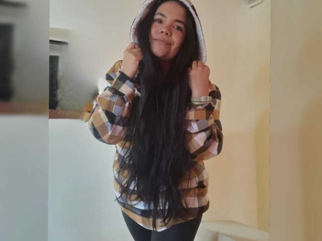 NoahJade with long black hair, smiling, wearing a brown and white plaid hoodie, holding the hood up with hands, standing indoors, looking cute and happy.