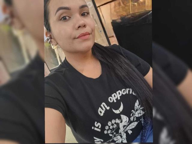 NoahJade has light brown skin, dark straight hair, small nose, and subtle smile. She wears a black T-shirt with a white floral design, showing her small tits.