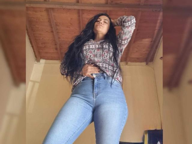 NoahJade with long black hair, wearing a patterned shirt and high-waisted blue jeans, standing with one hand on her hip, looking down confidently. Wooden ceiling above.