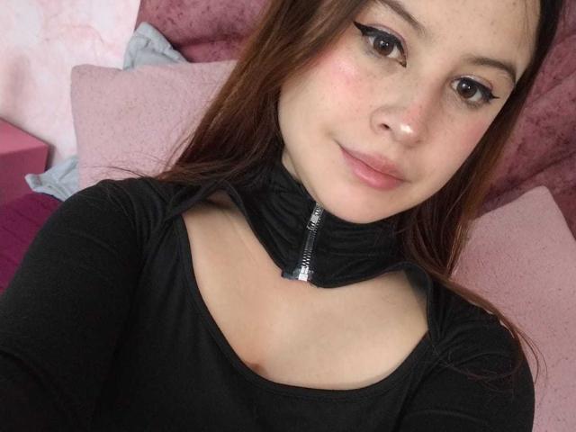 MelaniBecker has light skin, brown eyes with winged eyeliner, and brown hair. She's wearing a black zip-up top, showing her collarbones and smiling slightly. Boobs are not visible.