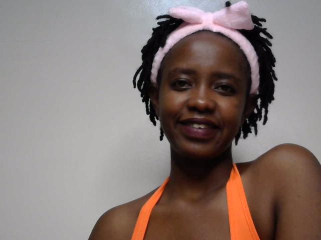 SweetHeidi with dark skin, black dreadlocks, pink headband with bow, orange halter top, smiling, showing her lips and teeth. Boobs not visible, focus on face and upper chest.