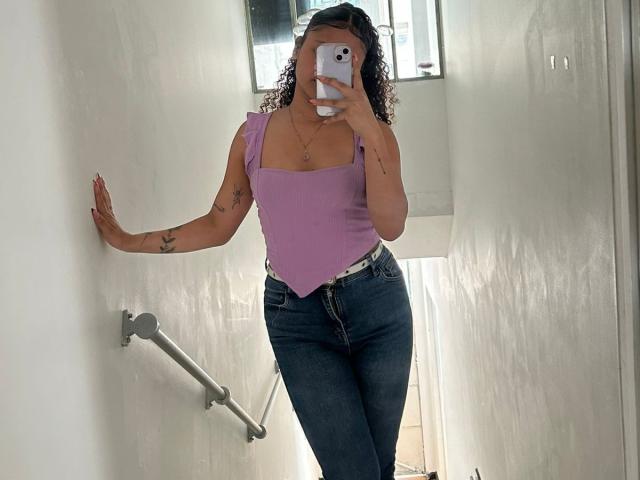 SofiaPassionne with curly hair, purple tank top, and high-waisted blue jeans, standing in a white bathroom, holding a phone, showing tattoos on her arms, and a small chest.