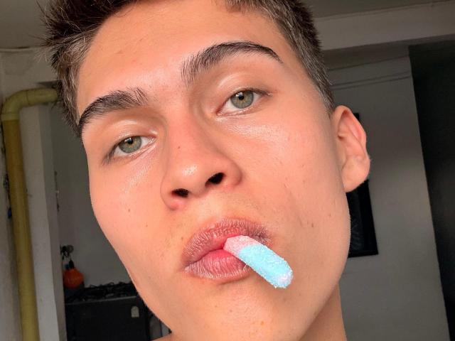 Bolaquesoo a une glace bleu clair entre ses lèvres gonflées et roses. On peut voir ses yeux verts, ses courts cheveux bruns et sa peau claire. Il regarde directement l'appareil photo..