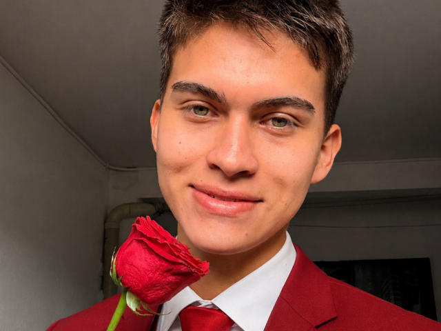 Bolaquesoo con una rosa roja en la boca, vistiendo un traje rojo, camisa blanca y corbata roja, sonriendo a la cámara, cabello castaño corto, ojos verdes, piel clara..