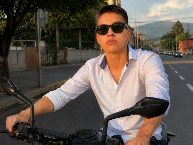 Bolaquesoo, wearing black sunglasses and a white shirt, sits on a motorcycle with hands on the handlebars, pouting his lips. Short brown hair, visible chest, sunny background.