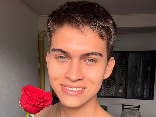 Bolaquesoo mit kurzen, braunen Haaren, grünen Augen und einem süßen Lächeln, der eine rote Rose vor seinem Gesicht hält. Er ist ohne Hemd, wodurch seine glatte, helle Brust zu sehen ist..