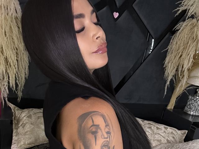 ValeryJhonston with long black hair, eyes closed, glossy pink lips, black sleeveless top, tattoo of a man's face on right arm, pink heart emoji in background.