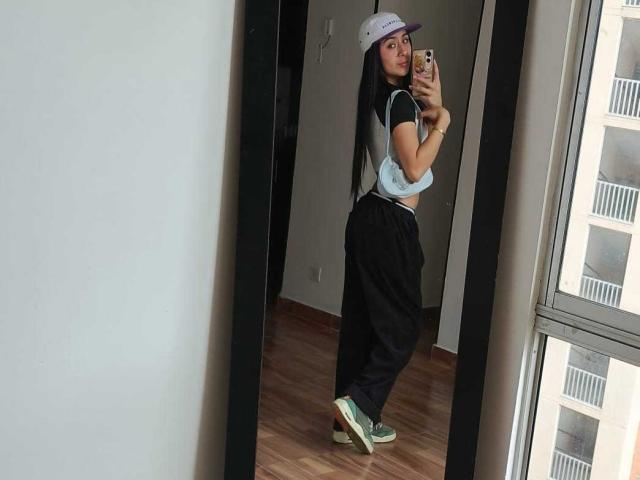 ChloeXk in a mirror selfie, wearing a white cap, black shirt, black pants, green and white sneakers, standing with her ass slightly raised, holding a phone.