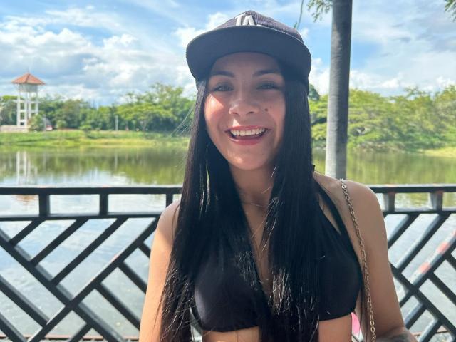 ChloeXk with long black hair, smiling, wearing a black cap and black bikini top, showing her small tits, standing in front of a metal railing by a reflective waterway.