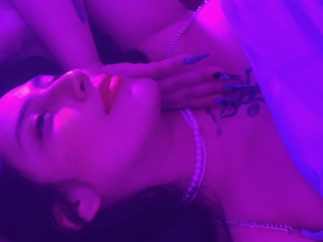 TatianaOnnel's face, purple-lit, with eyes closed, lips slightly parted, necklace, tattooed hand on chest, long blue nails, dark hair, white bed, relaxed, sensual.