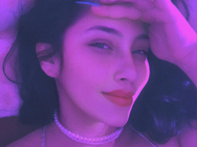 TatianaOnnel has purple-lit skin, dark wavy hair, and a pearl choker. She's winking with orange lipstick, holding her hair with one hand, and has blue nails.