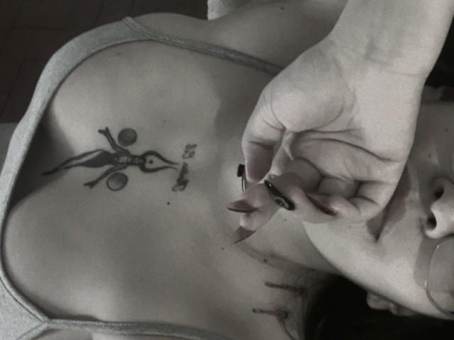 TatianaOnnel's chest has a black tattoo of a stick-figure woman with circles for eyes and a mouth, holding a small "no" symbol. She's using black nail polish.