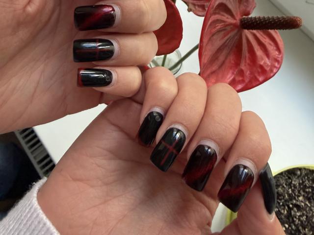 VikkiRi's hands with shiny black and red marbled nails, fingers slightly curled, holding a red leafy plant. Her skin is light, and she's wearing a white sleeve.