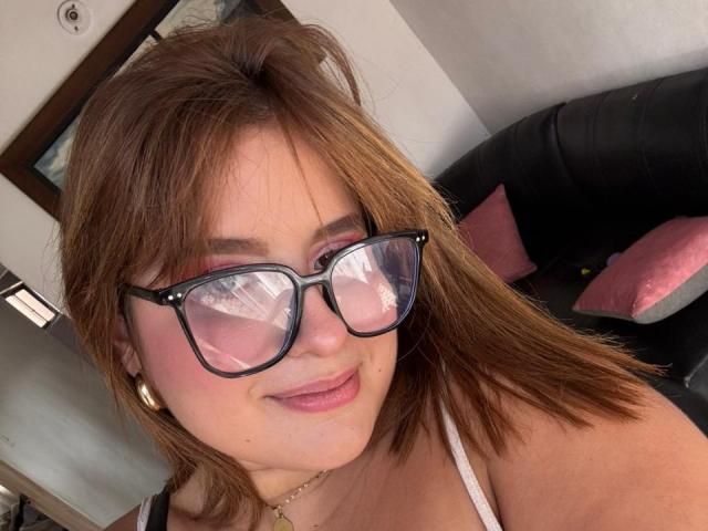 Ivy Oconner, usando óculos, com cabelo castanho, sombra rosa nos olhos, sorrindo, vestindo uma blusa branca, brincos dourados e um colar, com o reflexo de uma janela em seus óculos..