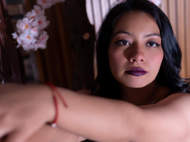 AshleyyBelle heeft donker haar, paarse lippenstift en kijkt met een serieuze uitdrukking in de camera. Haar armen zijn gekruist, en er is een rode armband te zien. Haar borsten zijn gedeeltelijk zichtbaar..