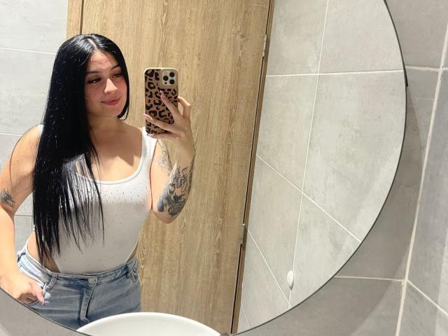 AnnyBentleii, com cabelos longos e pretos, usando uma camiseta branca que mostra os seios, com tatuagens nos braços, segurando uma capa de celular com estampa de leopardo, vestindo jeans de cintura alta, tirando uma selfie no espelho..
