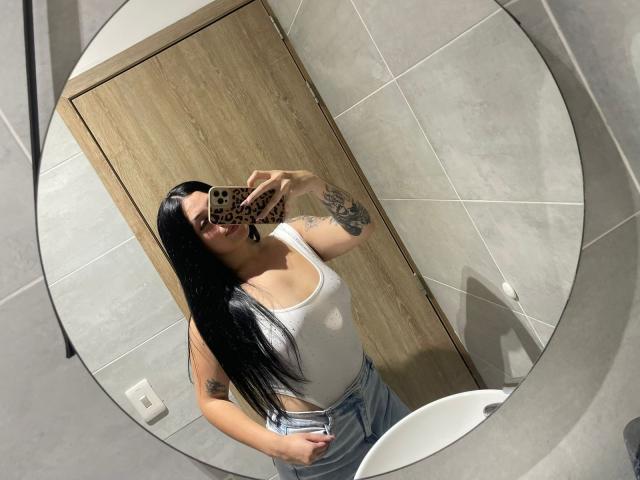 AnnyBentleii mit langen, schwarzen Haaren, die ein weißes Tanktop und blaue Jeans trägt und ihre Tattoos zeigt, macht ein Selfie vor einem Spiegel in einem gefliesten Badezimmer. Ihre Brüste sind sichtbar..