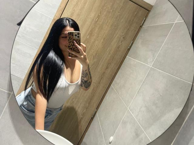 AnnyBentleii mit langen, schwarzen Haaren, die ein weißes Tanktop und hoch taillierte Jeans trägt, macht ein Selfie vor einem Spiegel, auf dem ihr tätowierter Arm zu sehen ist, und hält ein Handy mit Leopardenmuster..