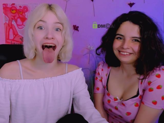 SabrinaMia with blonde hair and white off-shoulder top, tongue out, and another SabrinaMia with curly brown hair, pink strawberry-print top, smiling. Both have visible tits.