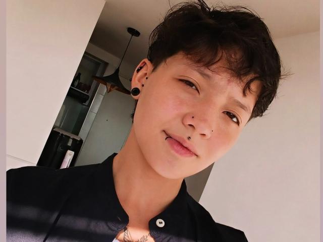 LissyLuxe has short, messy black hair, a cute nose ring, and is sticking her tongue out with black ear plugs. She's wearing a black shirt, looking adorable.