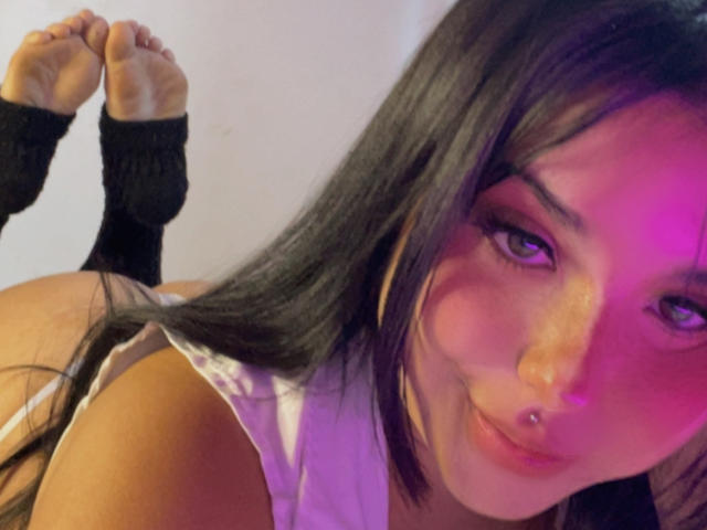 GraceClarke lying on her stomach, face close to camera, black hair, purple and pink lighting, wearing a pink top, black leg warmers, her bare ass and feet in the background.