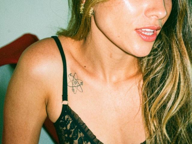 AdharaCooperr, met gebruinde huid, glanzende lippen en een zwarte kanten bh, toont een kleine tatoeage van een atoom op haar rechter schouder, met lang blond haar..