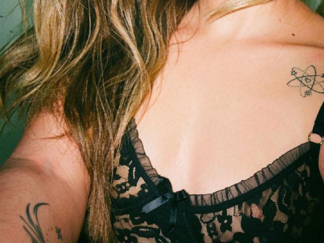 AdharaCooperr's chest with a black lace bra, showing a small tattoo of a double spiral on the right side and a tattoo on the left forearm. Blonde hair.