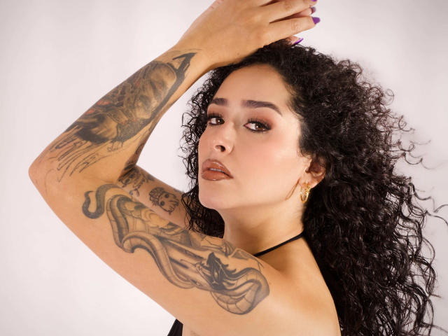 NatashaVelvet met krullend zwart haar, tatoeages op beide armen, paarse nagellak, gouden oorbellen, een zwarte haltertop, die omhoog kijkt met bruine lippenstift, en haar arm boven haar hoofd houdt..