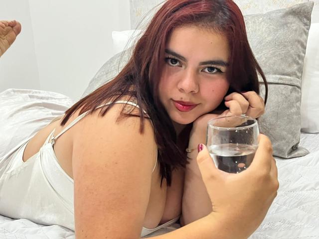 GeralGen with red hair, white satin dress, and red lipstick, holding a glass of water, lying on a bed, showing her tits and a slight smile, looking at the camera.
