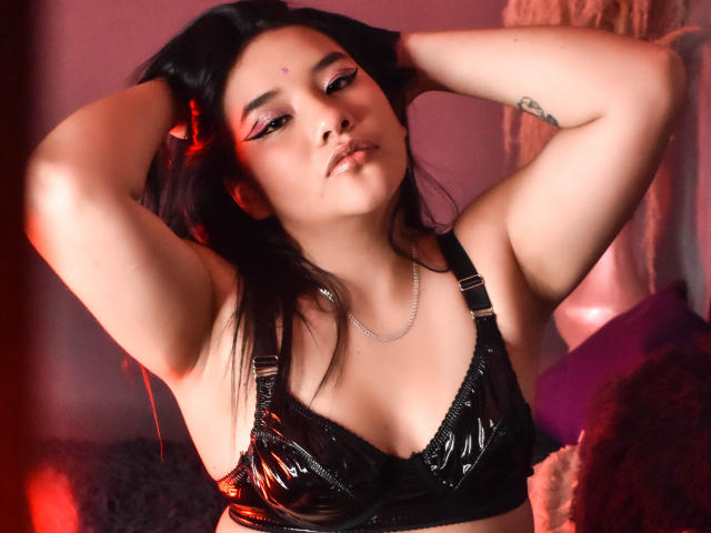CindyDecker met een zwarte, glanzende beha, handen in haar haar, een tatoeage op de rechterarm, rode oogmake-up, een lichte glimlach, een ketting, rode en roze verlichting, middelgrote borsten, Aziatisch, sexy..