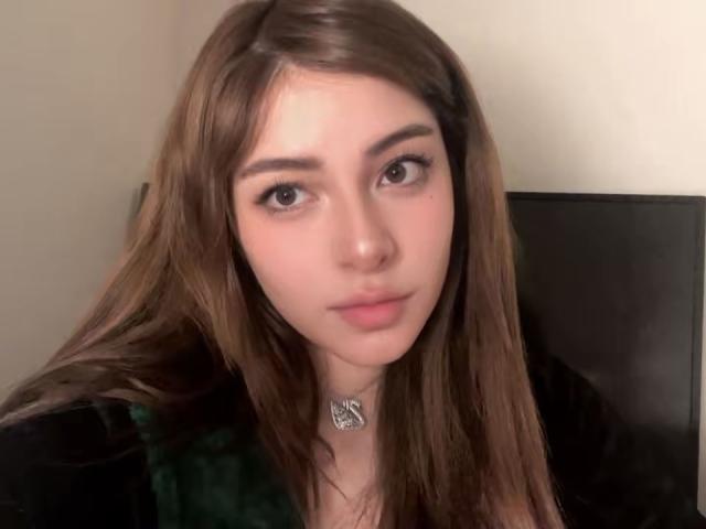 AngelinaNoAngel has long brown hair, brown eyes, and a slight smile. She's wearing a black top with a green plaid pattern and a silver necklace.